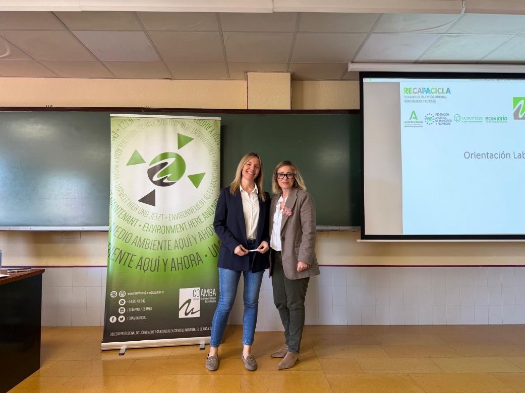 Dos personas de pie frente a una pantalla de proyector y una pancarta en un aula o sala de conferencias.