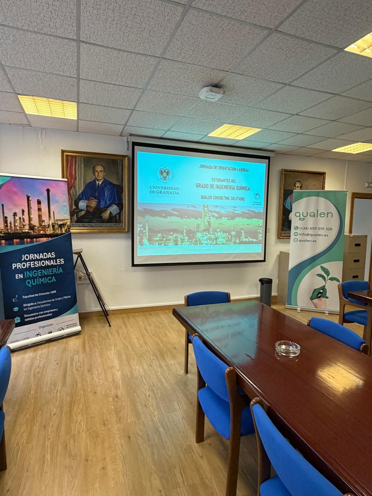 Sala de conferencias con pantalla de proyector, pancartas, retrato enmarcado y sillas azules alrededor de una mesa.