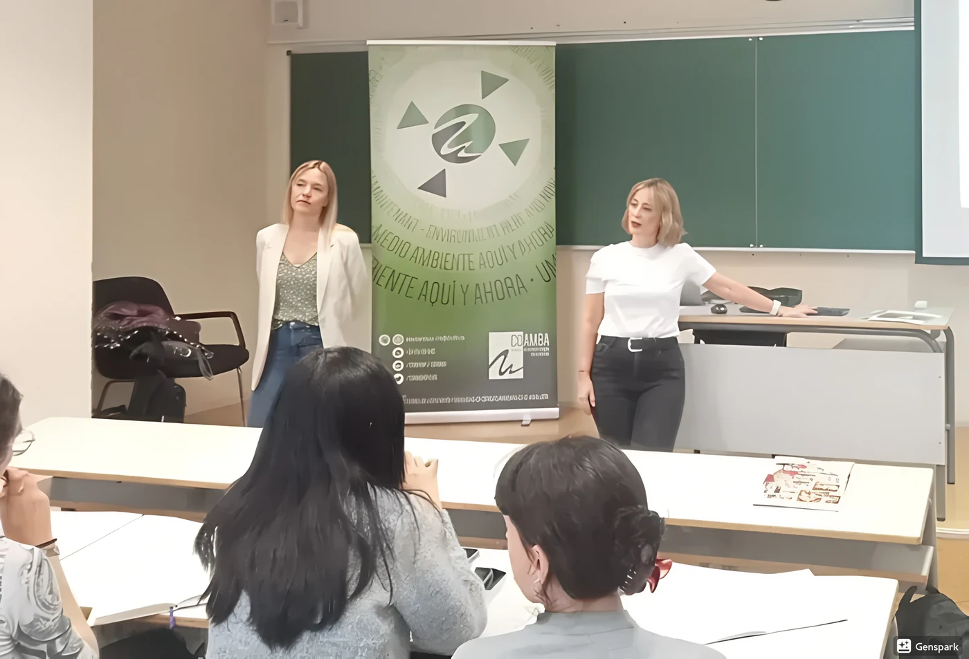 Dos mujeres haciendo una presentación al frente de un aula con una pancarta verde y una pizarra detrás.