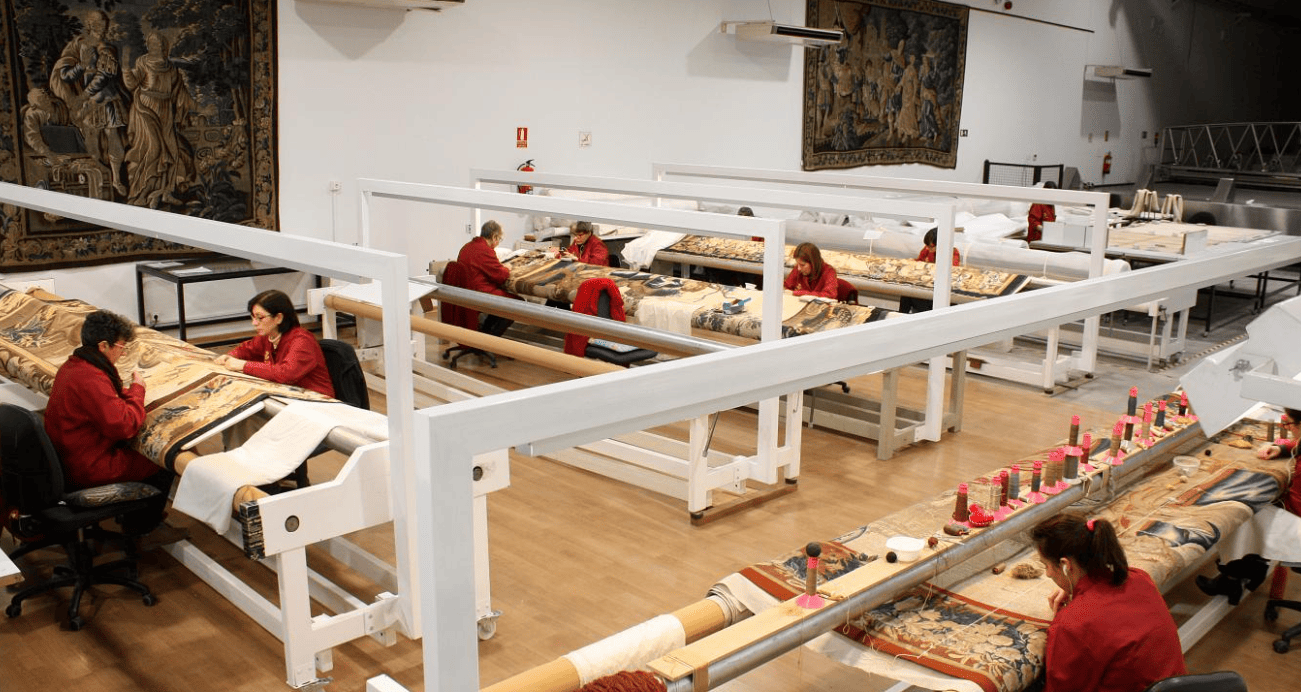 Personas tejiendo en grandes telares en un taller textil con bastidores blancos y materiales rojos.