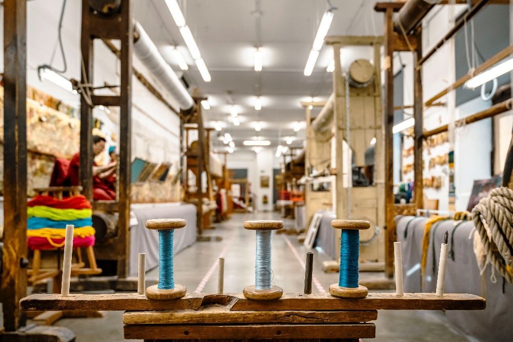Taller textil con telares, carretes de hilo y pilas de telas de colores en un largo pasillo.
